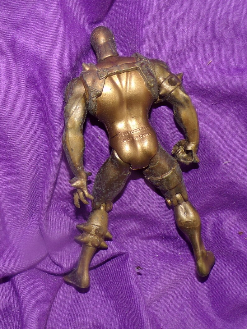 May include: A gold-colored plastic action figure with a spiky helmet and armor. The figure is posed with its arms raised and its legs spread apart. The figure is made of plastic and has a matte finish. The figure is standing on a purple surface.