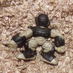 K&ouml;nnte beinhalten: Eine kleine, bewegliche Actionfigur in einer hockenden Pose. Die Figur ist haupts&auml;chlich grau mit schwarzer Panzerung auf Brust, Schultern, Ellbogen, Knien und F&uuml;&szlig;en. Der Helm ist schwarz. Gelbe Akzente sind an H&auml;nden und F&uuml;&szlig;en zu sehen.