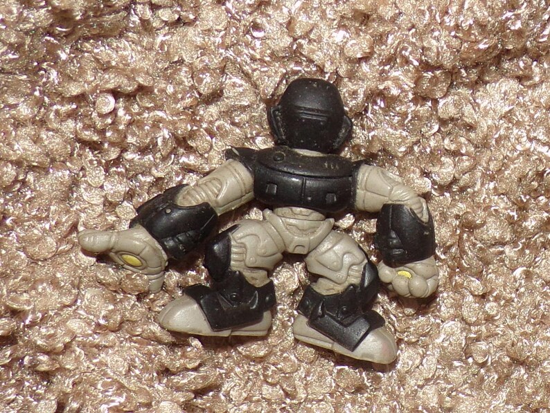 K&ouml;nnte beinhalten: Eine kleine, bewegliche Actionfigur in einer hockenden Pose. Die Figur ist haupts&auml;chlich grau mit schwarzer Panzerung auf Brust, Schultern, Ellbogen, Knien und F&uuml;&szlig;en. Der Helm ist schwarz. Gelbe Akzente sind an H&auml;nden und F&uuml;&szlig;en zu sehen.