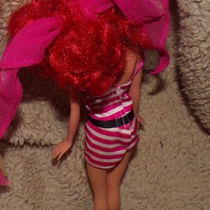 May include: A red-haired doll wearing a pink and white striped dress with a black belt. The doll has a pink ribbon in her hair.
