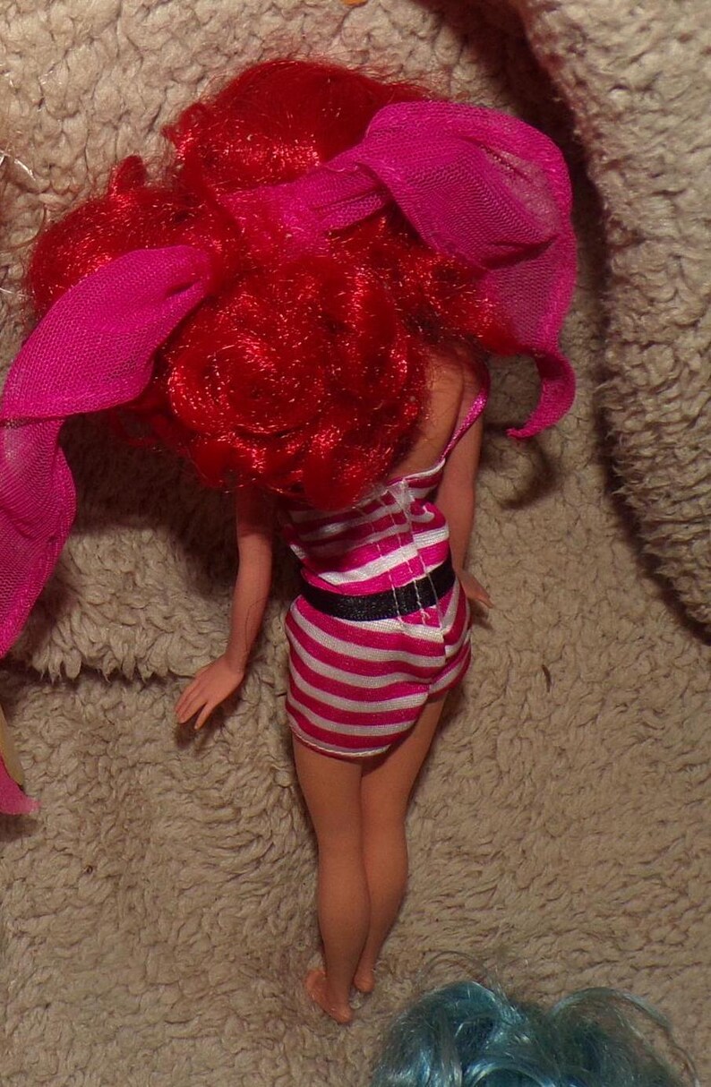 May include: A red-haired doll wearing a pink and white striped dress with a black belt. The doll has a pink ribbon in her hair.