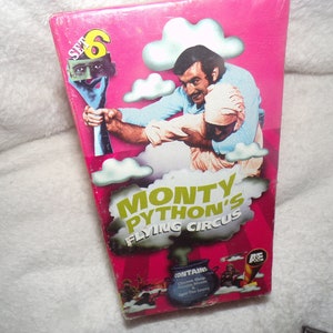 May include: A pink VHS tape case with a picture of a man holding another man in the air. The case has the text "Monty Python's Flying Circus" in yellow letters and "Set 6" in the top left corner.