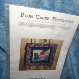 May include: A quilting pattern for a wall hanging titled "Purple Lady" by Plum Creek Patchwork, Rt 2 Box 95 Walnut Grove MN 56180. The pattern shows a framed quilt with a purple lady in a red hat standing in a doorway.