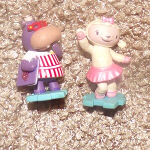 May include: Two small plastic figurines standing on light blue bases. One is a purple hippo wearing red glasses and a white and red striped shirt holding a purple book. The other is a cream-colored lamb wearing a pink skirt and bow.