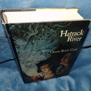 May include: A hardcover book titled "Hatrack River" by Orson Scott Card. The cover features a dark blue and green illustration of a young boy holding a glowing orb, with figures and a town in the background.