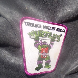 May include: A rectangular patch featuring a Teenage Mutant Ninja Turtle character. The patch has a white background with the words "TEENAGE MUTANT NINJA TURTLES" in black and green. The turtle is green and purple, with a red chest and a purple mask. The patch is outlined in pink.