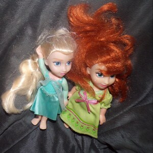 May include: Two small dolls, one with blonde hair in a side ponytail and a teal dress, the other with red hair and a green dress. Both dolls have blue eyes and are posed on a dark gray surface.