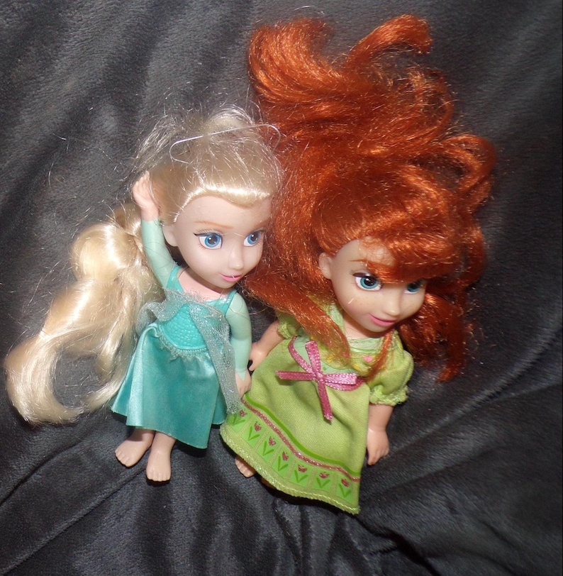 May include: Two small dolls, one with blonde hair in a side ponytail and a teal dress, the other with red hair and a green dress. Both dolls have blue eyes and are posed on a dark gray surface.