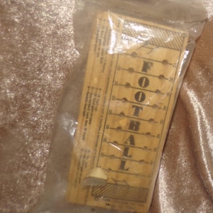 May include: A rectangular wooden board game titled FOOTBALL enclosed in a clear plastic bag. The board features a vertical grid with numerical markings and printed rules for gameplay, including yardage gains and point values for various football plays.