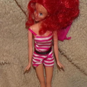 May include: A red-haired doll wearing a pink and white striped swimsuit with a black belt. The doll has blue eyes and is posed with her arms at her sides.