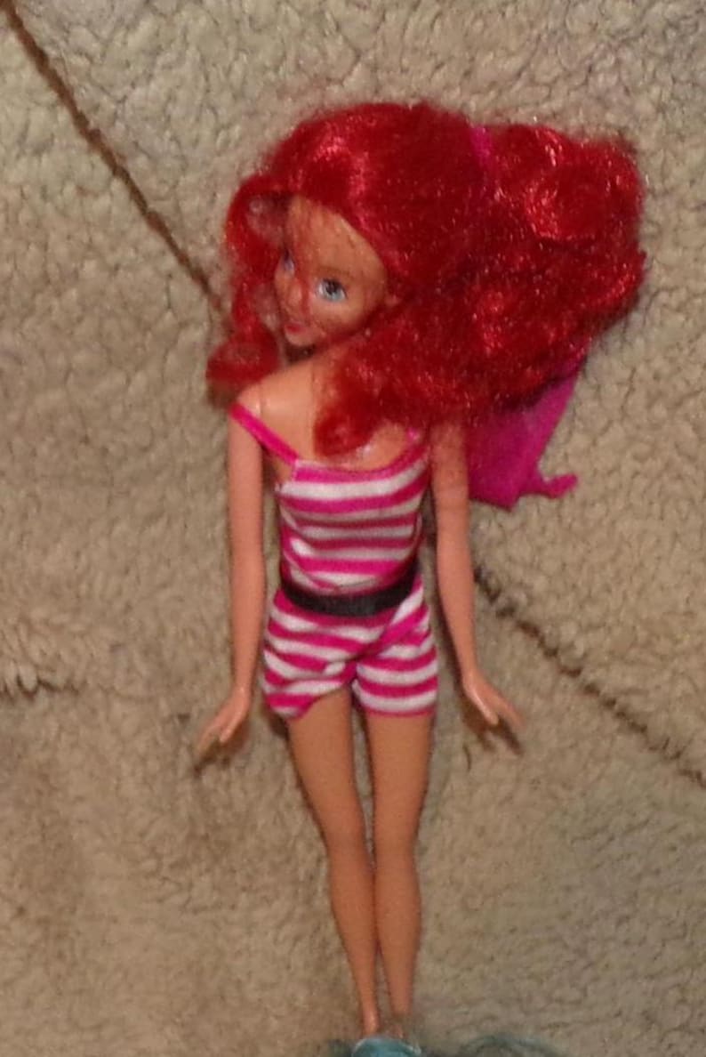 May include: A red-haired doll wearing a pink and white striped swimsuit with a black belt. The doll has blue eyes and is posed with her arms at her sides.