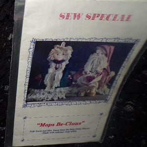May include: A printed paper with the text "SEW SPECIAL" in pink. The image features two folk art dolls, Santa and Mrs. Santa, with the text "Mops Be-Claus". The dolls are made with mop refills.