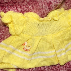 May include: A yellow knitted baby dress with white stripes and a duck applique. The dress has a ruffled neckline and sleeves. The fabric appears soft and lightweight, suitable for warm weather. The dress is likely for a baby or toddler.