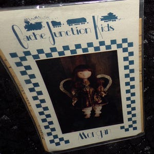 May include: A Cache Junction Kids doll package featuring a doll with a dark dress and white pants. The package has a blue and white checkered border and the name "Morgan" at the bottom. The Cache Junction Kids logo is at the top.