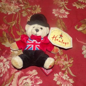 May include: A plush teddy bear wearing a black bowler hat, red jacket with a Union Jack design, black pants, and a red bow tie. The bear has a tag that says "Hamleys". The bear is sitting on a red floral background.