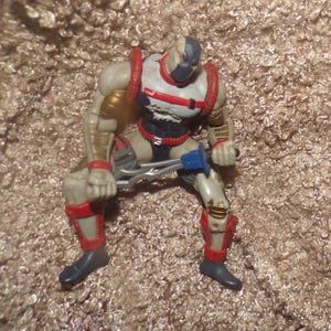 May include: A detailed action figure of a muscular character in a seated position, holding a tool. The figure has a white, red, and black color scheme with gold accents. The figure is wearing a mask and boots.