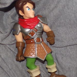 May include: A plastic action figure of a male character wearing brown armor, a red scarf, and green pants. The figure has brown hair and blue eyes.