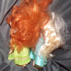 May include: Two dolls with long hair, one red and one blonde, are shown from the back. The doll on the left wears a light green dress with a floral pattern. The doll on the right wears a light blue dress.