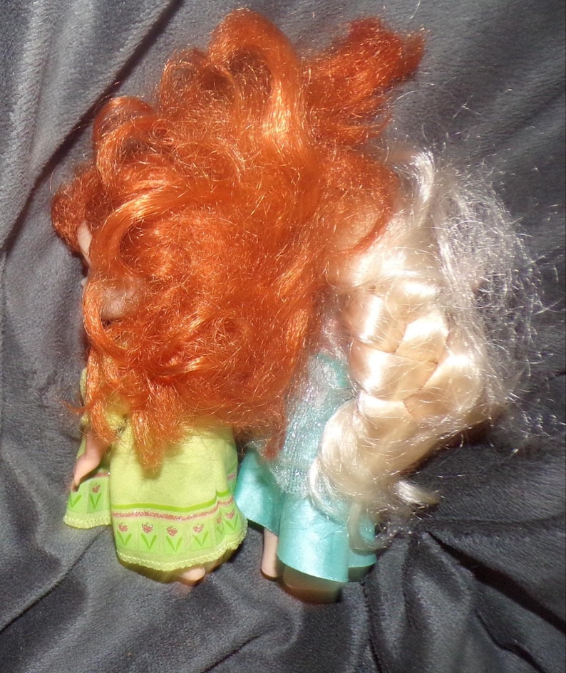 May include: Two dolls with long hair, one red and one blonde, are shown from the back. The doll on the left wears a light green dress with a floral pattern. The doll on the right wears a light blue dress.