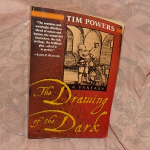 May include: A red book cover with the title "The Drawing of the Dark" in gold lettering. The author, Tim Powers, is printed in white lettering. A quote about the book is printed in white lettering on the left side of the cover. An illustration of a man in a tavern is printed on the right side of the cover.