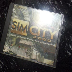 May include: A CD case for the computer game SimCity 3000. The case is silver and has a picture of a city on it. The text "SimCity 3000" is in gold letters and the text "Create and control your own urban empire" is in black letters.
