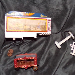 May include: Miniature circus toy set featuring a white and blue wagon with a painted scene, a red and gold wagon, and several wheels. Also included are two sets of white wheels. The toys are on a black surface.