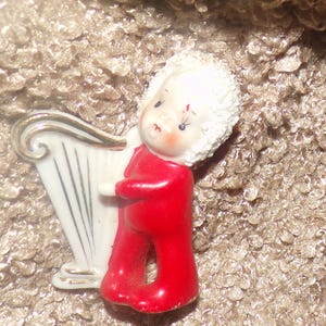 May include: A ceramic figurine of a small character with a textured white curly wig, wearing a solid red outfit, and holding a white harp with gold-colored trim. The character has painted facial features including dark eyes and rosy cheeks.
