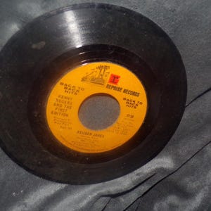 May include: A black vinyl record with a yellow and orange label. The label reads "BACK TO BACK HITS" and features the text "KENNY ROGERS AND THE FIRST EDITION". The record is a 7-inch single, a vintage music collectible.