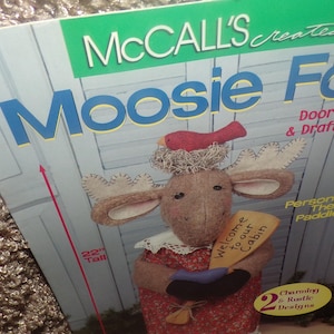 May include: A McCall's pattern for a Moosie Folk door-stopper and draft-dodger. The image shows a plush moose with a red bird on its head, holding a sign that says "Welcome to our Cabin." The moose is 22 inches tall.