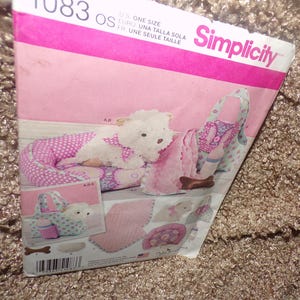 Pode incluir: Padrão de costura Simplicity para cama, transportador e acessórios para cães. O padrão apresenta um brinquedo de pelúcia para cães, uma cama de bolinhas rosa e um transportador floral. Inclui o texto "UMA TALLA SOLA".