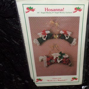 Może przedstawiać: Opakowanie z 40 cm aniołkiem królikiem i girlandą z aniołków królików. Opakowanie zawiera tekst "Hosanna!" i "Needle in a Haystack!!". Girlanda przedstawia trzy figurki królików ze skrzydłami anioła, kokardkami i wzorzystymi ubraniami.