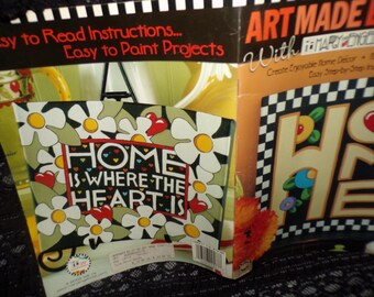May include: A paint-by-numbers kit featuring a colorful sign with the text "Home is where the heart is". The sign is surrounded by daisies and hearts. The kit is part of the Art Made Easy with Mary Engelbreit collection.