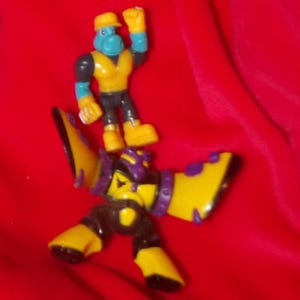 May include: Two action figures on a red background. One figure is a blue-skinned humanoid wearing a yellow vest, hat, and boots, with black pants and arms. The other figure is a yellow and purple robot with black accents.