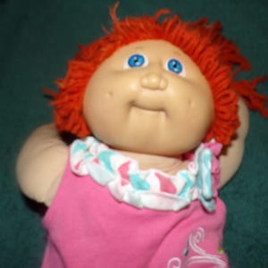 May include: A vintage doll with a fair complexion, blue eyes, and bright orange yarn hair. The doll wears a pink top with a ruffled collar featuring a pattern of white, light blue, and pink. The doll's arms are visible.
