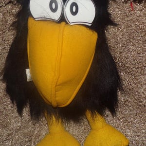 May include: A plush toy bird with black faux fur, a large yellow fabric beak, and two oversized white plastic eyes with black pupils. The bird has two yellow fabric feet and a small white tag attached to the side of its black feathered body.