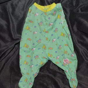 May include: A light green baby sleeper with a yellow collar and footed bottoms. The sleeper is patterned with yellow rocking horses, teddy bears, and other baby-themed graphics. The fabric appears to be cotton.