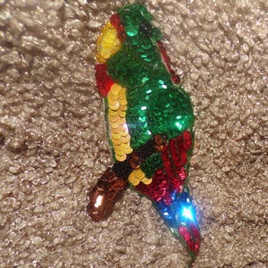 May include: A colorful parrot-shaped sequin embellishment. The bird features green, red, yellow, and blue sequins. The parrot's body is primarily green, with red and yellow accents. The tail is blue, and the beak is brown.
