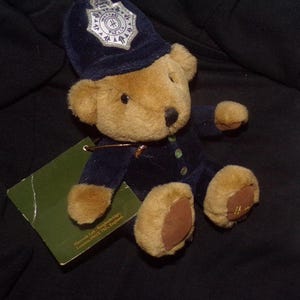 May include: A plush teddy bear wearing a navy blue police uniform and hat. The bear has tan fur, brown paws, and a badge on the hat. A green tag is attached to the bear.