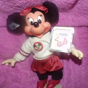 Puede incluir: Una mu&ntilde;eca de peluche Minnie Mouse roja y blanca con una falda roja y una camisa blanca con una raya roja y blanca en la manga. La camisa tiene un c&iacute;rculo rojo con una imagen en blanco y negro de Mickey Mouse dentro. La mu&ntilde;eca sostiene una etiqueta que dice "Nodda - Feliz cumplea&ntilde;os".