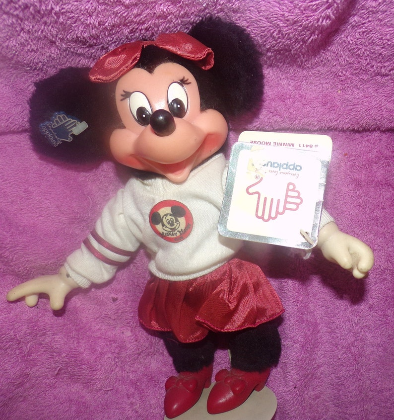 Puede incluir: Una mu&ntilde;eca de peluche Minnie Mouse roja y blanca con una falda roja y una camisa blanca con una raya roja y blanca en la manga. La camisa tiene un c&iacute;rculo rojo con una imagen en blanco y negro de Mickey Mouse dentro. La mu&ntilde;eca sostiene una etiqueta que dice "Nodda - Feliz cumplea&ntilde;os".
