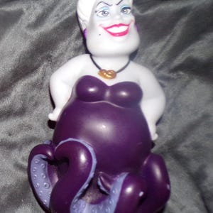 May include: A figurine of Ursula, the sea witch from Disney's The Little Mermaid. The figure has a white head with white hair, a purple body, and purple tentacles. She wears a black necklace with a gold pendant and a purple strapless dress.