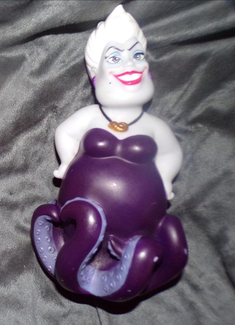 May include: A figurine of Ursula, the sea witch from Disney's The Little Mermaid. The figure has a white head with white hair, a purple body, and purple tentacles. She wears a black necklace with a gold pendant and a purple strapless dress.
