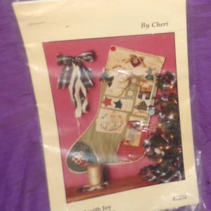 May include: A colorful patchwork stocking with a red, white, and blue bow. The stocking is filled with small, colorful squares and has a white background. The text "Filled with Joy" and "Quilting Pattern Packet" is printed below the stocking. The text "By Cheri" is printed in the upper right corner of the image.