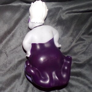 May include: A figurine of a character with white hair, a pale body, and a dark purple dress. The figurine is posed with its back to the viewer, showcasing the details of the character's hair and dress. The figurine is on a grey surface.