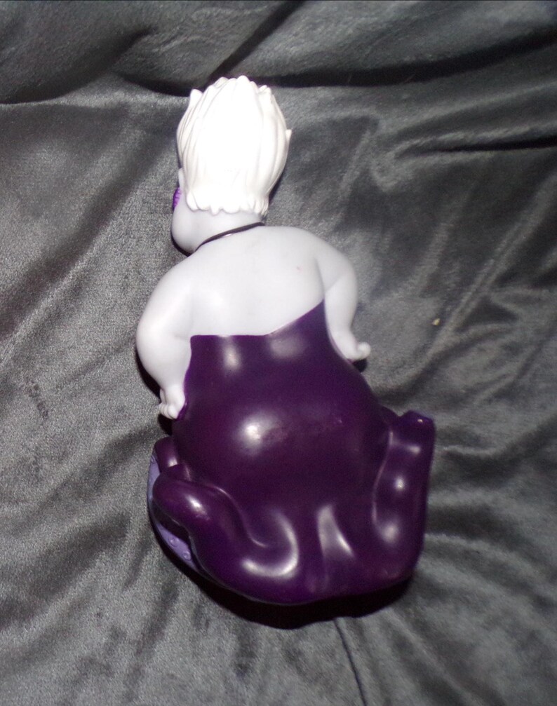 May include: A figurine of a character with white hair, a pale body, and a dark purple dress. The figurine is posed with its back to the viewer, showcasing the details of the character's hair and dress. The figurine is on a grey surface.