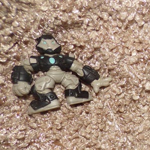 K&ouml;nnte beinhalten: Eine kleine Actionfigur eines roboterartigen Charakters. Die Figur ist haupts&auml;chlich grau mit schwarzen Akzenten auf der R&uuml;stung und den Stiefeln. Ein t&uuml;rkis farbener Kreis befindet sich auf der Brust. Die Figur ist auf einer strukturierten, beigefarbenen Oberfl&auml;che platziert.