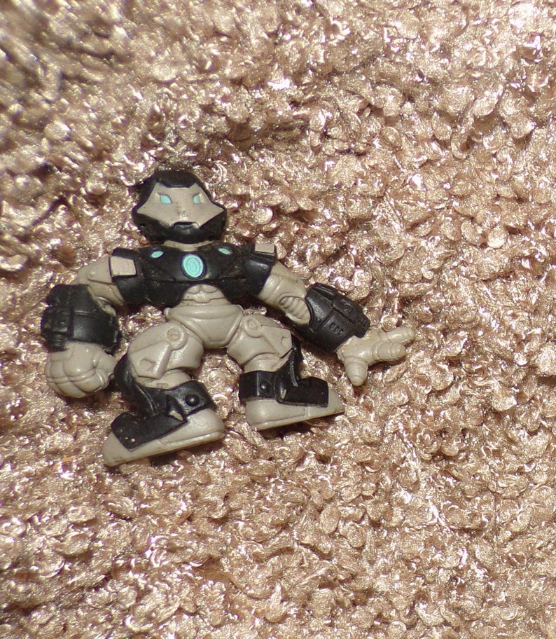 K&ouml;nnte beinhalten: Eine kleine Actionfigur eines roboterartigen Charakters. Die Figur ist haupts&auml;chlich grau mit schwarzen Akzenten auf der R&uuml;stung und den Stiefeln. Ein t&uuml;rkis farbener Kreis befindet sich auf der Brust. Die Figur ist auf einer strukturierten, beigefarbenen Oberfl&auml;che platziert.