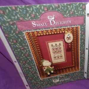 May include: A cross stitch pattern featuring a winter scene with a snowman and the text "Be Merry". The pattern is titled "Small Delights" and is by "Just Nan".