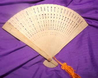 May include: A wooden folding fan with a decorative pattern and an orange tassel. The fan is closed and has a light brown color.
