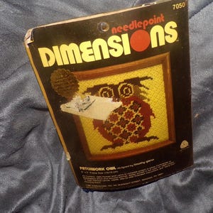 May include: A vintage Dimensions needlepoint kit featuring a patchwork owl design. The kit includes a framed needlepoint with a yellow background and a brown and red owl. The packaging is black with yellow text and the Dimensions logo.